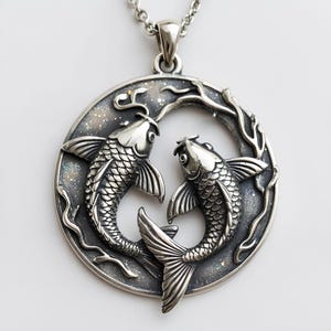 May include: A silver pendant necklace featuring two koi fish in a circular design. The fish are detailed with scales and fins, set against a textured, glittery background. The pendant hangs from a delicate silver chain.