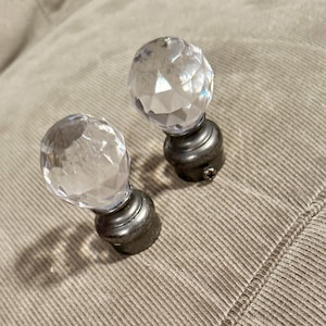 Könnte beinhalten: Zwei dekorative Gardinenstangen-Endstücke. Jedes Endstück hat einen facettierten, klaren Kristallkugel auf einem dunklen Metallsockel. Die Sockel haben ein dekoratives, abgerundetes Design. Die Endstücke liegen auf einem hellbraunen Stoff.