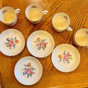 Può includere: Un set di tazze e piattini da tè vintage. Le tazze e i piattini sono bianchi con un motivo floreale di rose rosa, gialle e blu. I bordi sono rifiniti in oro. Il set comprende tre tazze e cinque piattini.