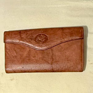 May include: A brown leather wallet with a flap closure. The wallet has a textured surface and a decorative emblem on the front. The wallet is rectangular and appears to be vintage.