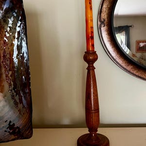 Può includere: Un candelabro in legno con un design tornito, che tiene una candela rossa e arancione. Il candelabro è posto su una superficie bianca. Uno specchio decorativo e un'opera d'arte sono visibili sullo sfondo. Il candelabro è alto circa 30 cm.