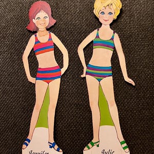 May include: Two vintage paper dolls, Jennifer and Julie, each wearing a striped bikini. Jennifer has red hair and blue shoes, while Julie has blonde hair and green shoes. The dolls are on a green and white base.