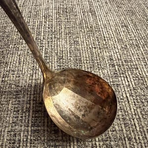 Puede incluir: Un cucharón de metal plateado con un cuenco profundo y redondeado y un mango largo y delgado. La superficie del metal muestra signos de oxidación y deslustre naturales, creando una pátina oscura y moteada en el interior del cuenco.