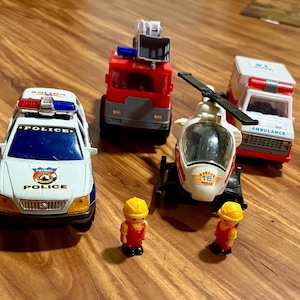 May include: A selection of toy emergency service vehicles: a police car, fire engine, ambulance, and helicopter. The police car is white and blue. The fire engine is red. The helicopter is white and red. Two toy construction workers are also present.