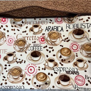 Peut inclure: Une pochette zippée crème avec un motif sur le thème du café. Le motif comprend des tasses à café, des grains et du texte tel que "Espresso Beans", "Latte" et "Arabica". La bordure supérieure est en tissu marron avec un motif tourbillonnant.