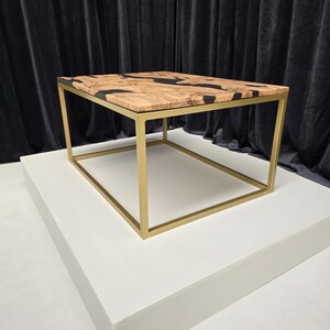 May include: A square coffee table with a gold metal frame and a wooden top with black epoxy resin. The table is on a white platform.