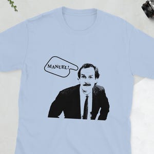 Puede incluir: Una camiseta azul claro con una imagen en blanco y negro de un hombre con bigote y una burbuja de diálogo que dice "¡MANUEL!"