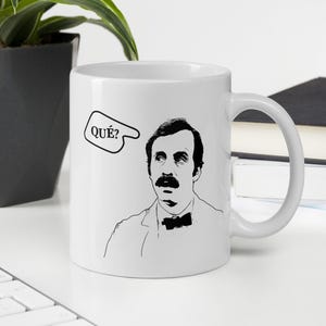 Può includere: Tazza in ceramica bianca con un'illustrazione in bianco e nero di un uomo con i baffi e la parola "QUE?" in un fumetto.