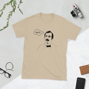 Puede incluir: Una camiseta beige con una ilustración en blanco y negro de un hombre con bigote y la palabra "QUE?" en un globo de diálogo.