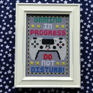 May include: Framed cross-stitch art with a gaming theme. The design features a white game controller with the text "GAMING IN PROGRESS DO NOT DISTURB!" in various colors. The frame is white, and the background is a navy blue fabric with white stars.