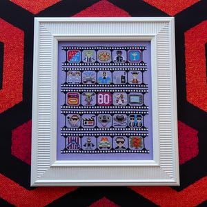 Puede incluir: Obra de arte en punto de cruz enmarcada que representa una tira de película con iconos temáticos de los años 80 y el número "80". La obra está sobre un fondo lila y enmarcada en un marco blanco texturizado. El fondo presenta un patrón geométrico rojo y negro.
