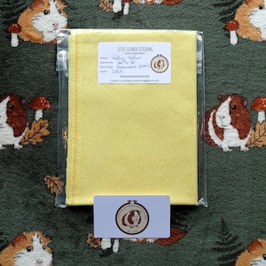 May include: A package containing a yellow fabric square, labelled "Molly Yellow," measuring 66 x 46 cm. The fabric is in a clear plastic bag, with a small business card at the bottom. The background features a green fabric with a guinea pig pattern.