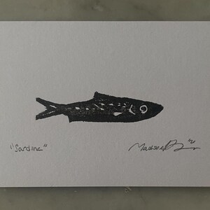 May include: A black ink block print of a sardine on a white rectangular card. The word Sardine is written in pencil on the bottom left, and a handwritten signature with the year 25 is located on the bottom right of the white paper surface.