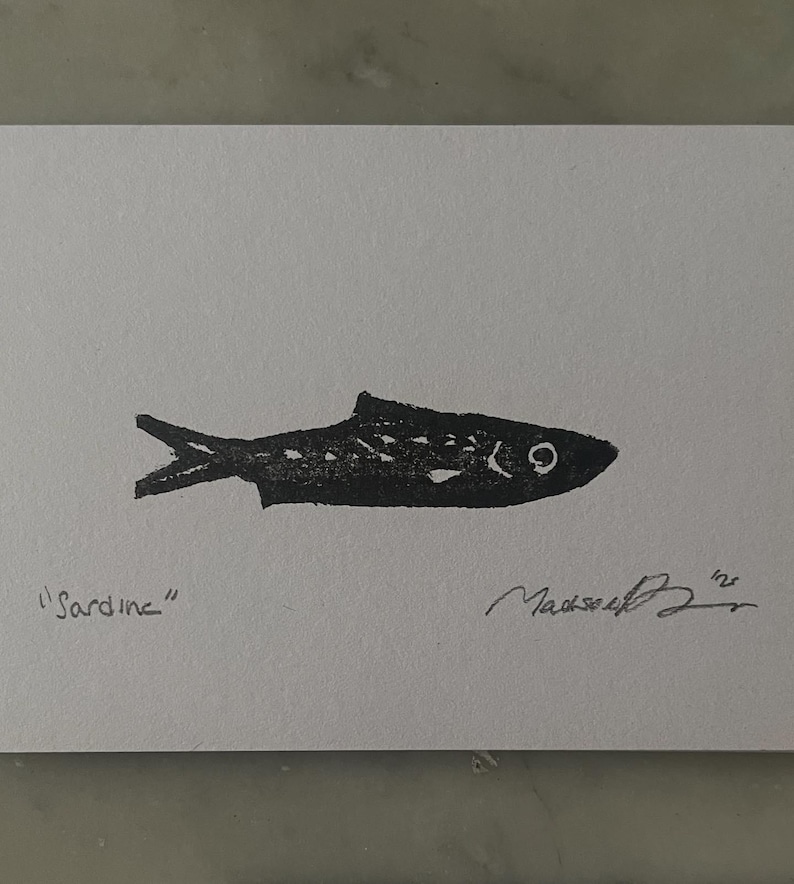 May include: A black ink block print of a sardine on a white rectangular card. The word Sardine is written in pencil on the bottom left, and a handwritten signature with the year 25 is located on the bottom right of the white paper surface.