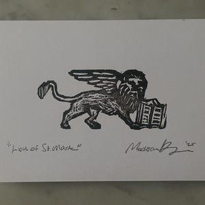 May include: A black ink block print on white paper featuring the Lion of St. Mark with wings and a book. The text "Lion of St. Mark" is written in the bottom left corner, and a handwritten signature with the year '25 is in the bottom right.