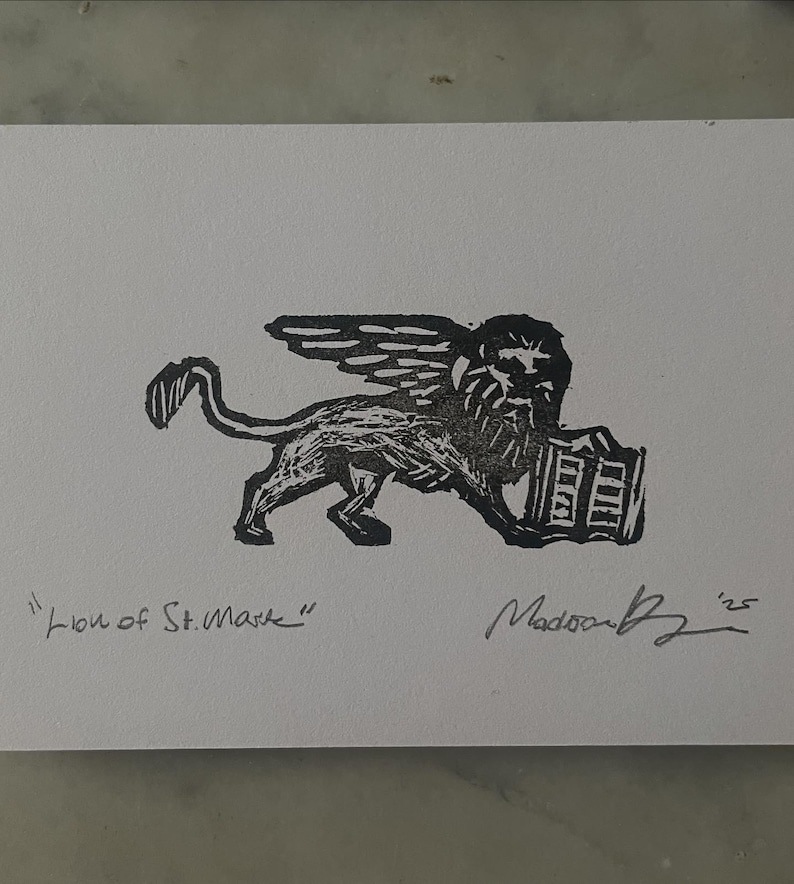 May include: A black ink block print on white paper featuring the Lion of St. Mark with wings and a book. The text "Lion of St. Mark" is written in the bottom left corner, and a handwritten signature with the year '25 is in the bottom right.