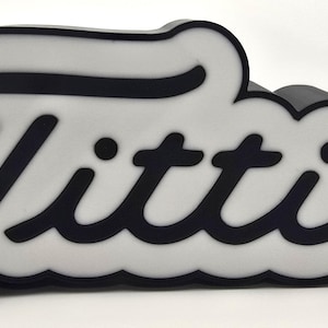 May include: A black and white sign with the word "Titties" in a bold, cursive font. The letters are black with a white outline, set against a white background. The sign has a rounded, cloud-like shape.