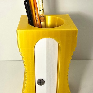May include: A yellow plastic pencil holder with a built-in pencil sharpener. The holder has a cylindrical opening at the top and a white sharpener compartment with a screw. Four pencils are in the holder.