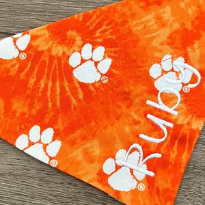 May include: Orange and white tie-dye dog bandana with white paw prints and the word "Rubys" embroidered in white.