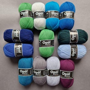 May include: A collection of Opal brand yarn skeins in various colors, including green, white, blue, teal, gray, and purple. Each skein is labeled with the brand name and product information. The yarn is arranged on a neutral gray surface.