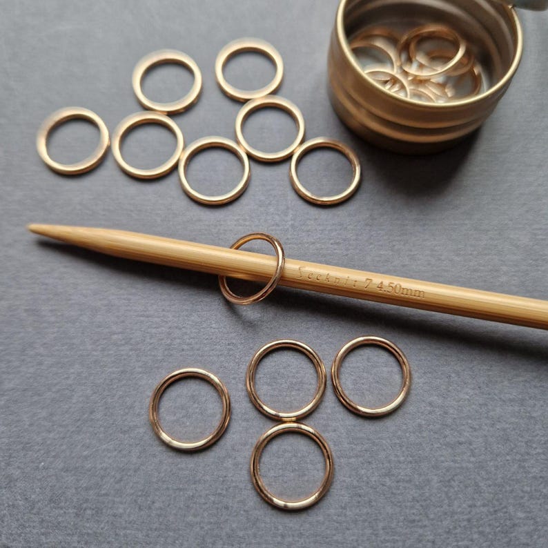 May include: A collection of gold-colored stitch markers, some scattered and others in a small container. A bamboo knitting needle with the text "Seeknit 7 4.50mm" is also present, with a stitch marker on it. The markers are used for knitting.