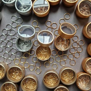 May include: An overhead shot displays small metal rings in various colors, including silver and gold. The rings are contained in small, round tins with clear lids. Some rings are scattered around the tins on a gray surface. The image suggests craft supplies or jewelry findings.