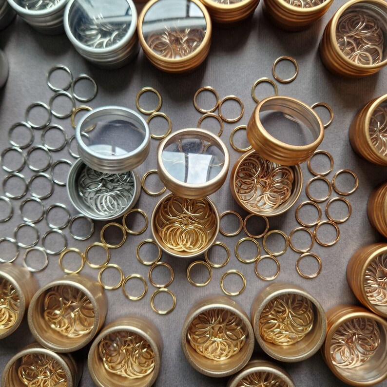 May include: An overhead shot displays small metal rings in various colors, including silver and gold. The rings are contained in small, round tins with clear lids. Some rings are scattered around the tins on a gray surface. The image suggests craft supplies or jewelry findings.