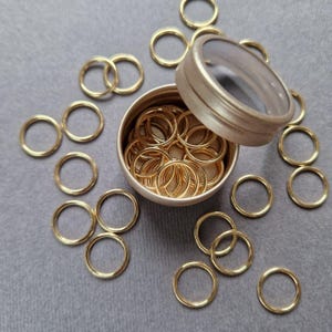 May include: A collection of gold-colored metal rings, some interlocked, scattered on a gray surface. A small, round, lidded container holds a larger quantity of the rings. The rings appear to be of uniform size and thickness.