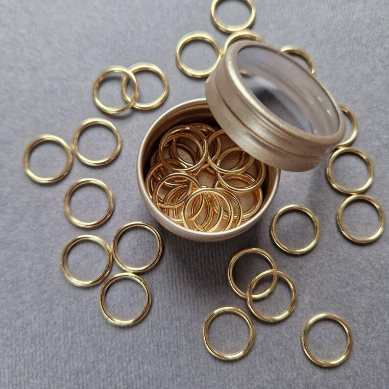May include: A collection of gold-colored metal rings, some interlocked, scattered on a gray surface. A small, round, lidded container holds a larger quantity of the rings. The rings appear to be of uniform size and thickness.