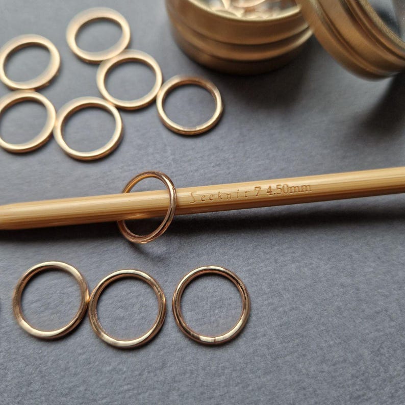 May include: A collection of gold-colored metal stitch markers and a bamboo knitting needle. The needle has the text "Seeknit: 7 4.50mm" printed on it. The stitch markers are circular and lie on a gray surface.