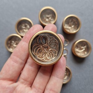 May include: A hand holding a gold-colored container filled with small gold rings. Several other similar containers are arranged around the main one. The containers have a decorative design on the outside.