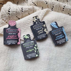 Puede incluir: Cuatro pequeños sobres negros de concentrado de lavandería Eucalan, cada uno con un aroma diferente: pomelo, jazmín, eucalipto y sin perfume. Los sobres están sobre una superficie de punto color crema. Cada sobre es de 5 ml.