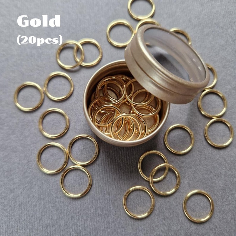 May include: Gold-colored metal rings, displayed both inside and around a small, round, lidded container. The text "Gold (20pcs)" is visible in the upper left corner. The rings are evenly spaced on a gray surface.