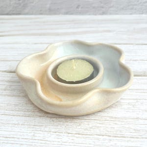 May include: A ceramic candle holder with a wavy, flower-like rim and a smooth, off-white glaze. The centre features a circular indentation holding a small, round, pale yellow tea light candle with a visible wick in the centre of the wax.