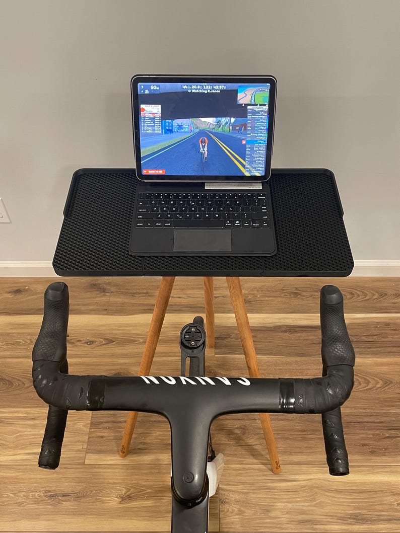 May include: A black indoor cycling setup with a tablet displaying a cycling simulation. The tablet is on a black stand with a keyboard, supported by a wooden tripod. The handlebars are black with the brand name "CANYON" in white.