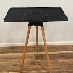 May include: A black, square table with a textured surface and a central circular cutout. The table is supported by three wooden legs. The table's dimensions are approximately 24 inches wide and 24 inches long.