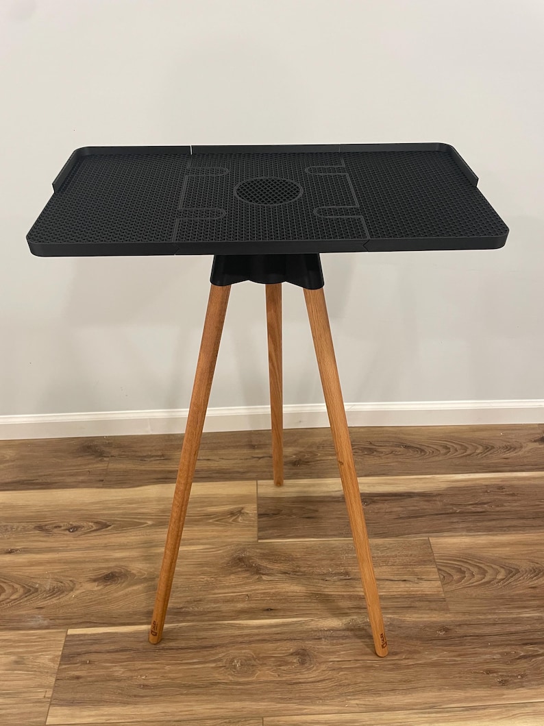 May include: A black, square table with a textured surface and a central circular cutout. The table is supported by three wooden legs. The table's dimensions are approximately 24 inches wide and 24 inches long.