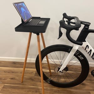 May include: A black tablet stand with wooden legs supports a tablet and keyboard. A white and black road bike is positioned next to the stand. The bike has black wheels and the word "Canyon" is visible on the handlebars.