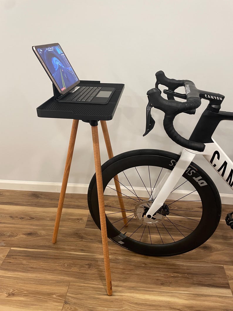 May include: A black tablet stand with wooden legs supports a tablet and keyboard. A white and black road bike is positioned next to the stand. The bike has black wheels and the word "Canyon" is visible on the handlebars.