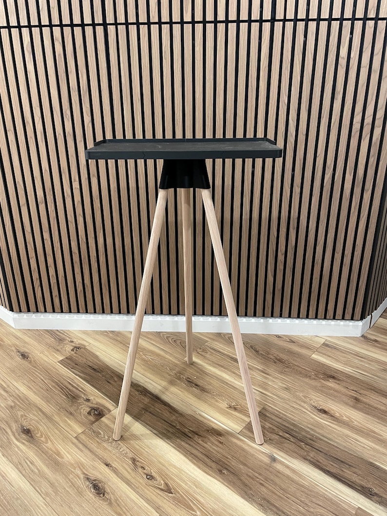 May include: A modern side table with a black square top and three light-colored wooden legs. The legs are angled outward for stability. The table is designed for indoor use and has a minimalist aesthetic.