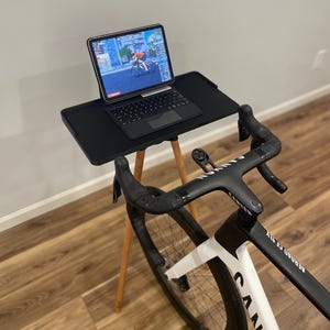 May include: A black and white Canyon Aeroad CF SLX bicycle with a black handlebar is set up for indoor cycling. A black laptop is on a small black table with wooden legs. The laptop screen displays a cycling simulation.