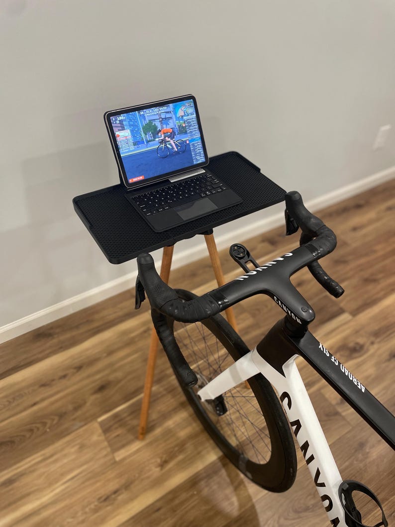 May include: A black and white Canyon Aeroad CF SLX bicycle with a black handlebar is set up for indoor cycling. A black laptop is on a small black table with wooden legs. The laptop screen displays a cycling simulation.