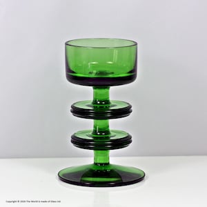 May include: A green glass candle holder with a circular base, a stem with two circular accents, and a bowl-shaped top. The glass has a dark green rim and accents. The design is simple and elegant.
