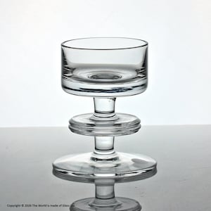 May include: A clear glass candle holder with a wide, shallow bowl and a tiered stem. The base is a flat circle, and the stem features two stacked, ridged discs. The candle holder is designed to hold a small candle.
