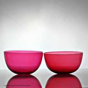 Puede incluir: Dos cuencos de vidrio magenta redondos a juego. Los cuencos tienen un acabado liso y brillante y un borde oscuro. Los cuencos están sobre una superficie reflectante, con sus reflejos visibles debajo. El fondo es blanco liso.