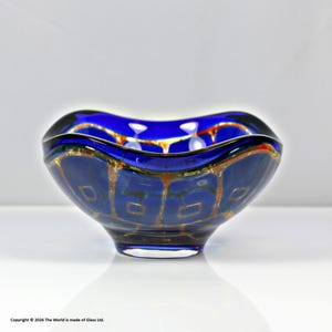 Può includere: Una ciotola di vetro blu vibrante con una forma e un design unici. La ciotola presenta un esterno blu scuro con motivi geometrici color oro. Il bordo ha un design ondulato con un bordo nero. Il copyright indica "Copyright © 2026 The World is made of Glass Ltd."