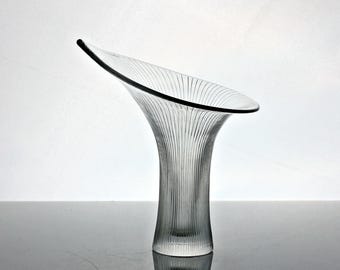 May include: A clear glass vase with a flared, asymmetrical opening and a narrow base. The surface features vertical, etched lines that extend from the bottom to the rim. The text Copyright 2026 The World is made of Glass Ltd. is visible.