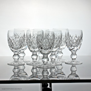 May include: A set of seven clear crystal glasses featuring a diamond-cut pattern on the bowl, a short stem with a spherical node, and a circular base. The glasses are arranged in a group, reflecting light through their intricate faceted surfaces.
