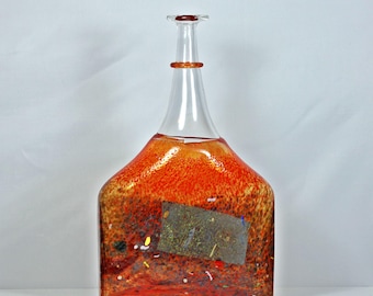May include: A decorative glass bottle with an orange and brown speckled design. The bottle has a wide base and a narrow neck with a red stopper. The bottle's design includes red, yellow, and brown hues. The bottle is likely for decorative purposes.