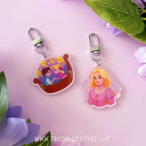 May include: Two clear acrylic keychains with silver lobster clasps. One keychain features a cartoon illustration of a couple in a wooden boat with floating lanterns. The other keychain shows a cartoon portrait of a blonde character in a pink dress.